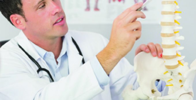 Chiropractor examining a patient’s lower back to assess herniated disc symptoms, with spine anatomy diagram in the background in Grand Rapids, MI