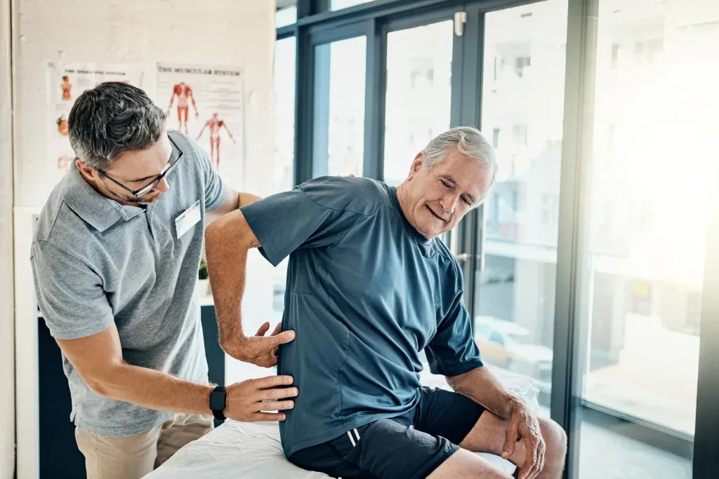 A healthcare professional is assisting a person seated on an examination table by examining their lower back in a well-lit clinical setting. This image could represent physical therapy or chiropractic care services available in Grand Rapids, MI.