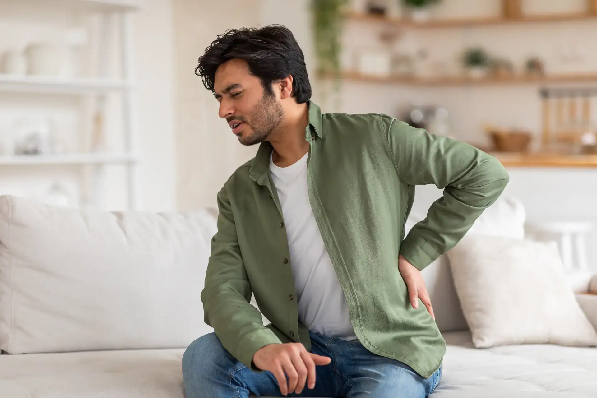 A middle-aged man is shown clutching his lower back in pain while standing indoors. This image effectively depicts the need for back pain relief services in Grand Rapids, MI.