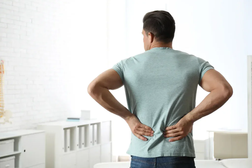 A man stands outdoors on a sunny day in Grand Rapids, MI, wearing casual clothing and holding his lower back in discomfort. This image effectively illustrates the issue of back pain and can be used to promote physical therapy services in the area.
