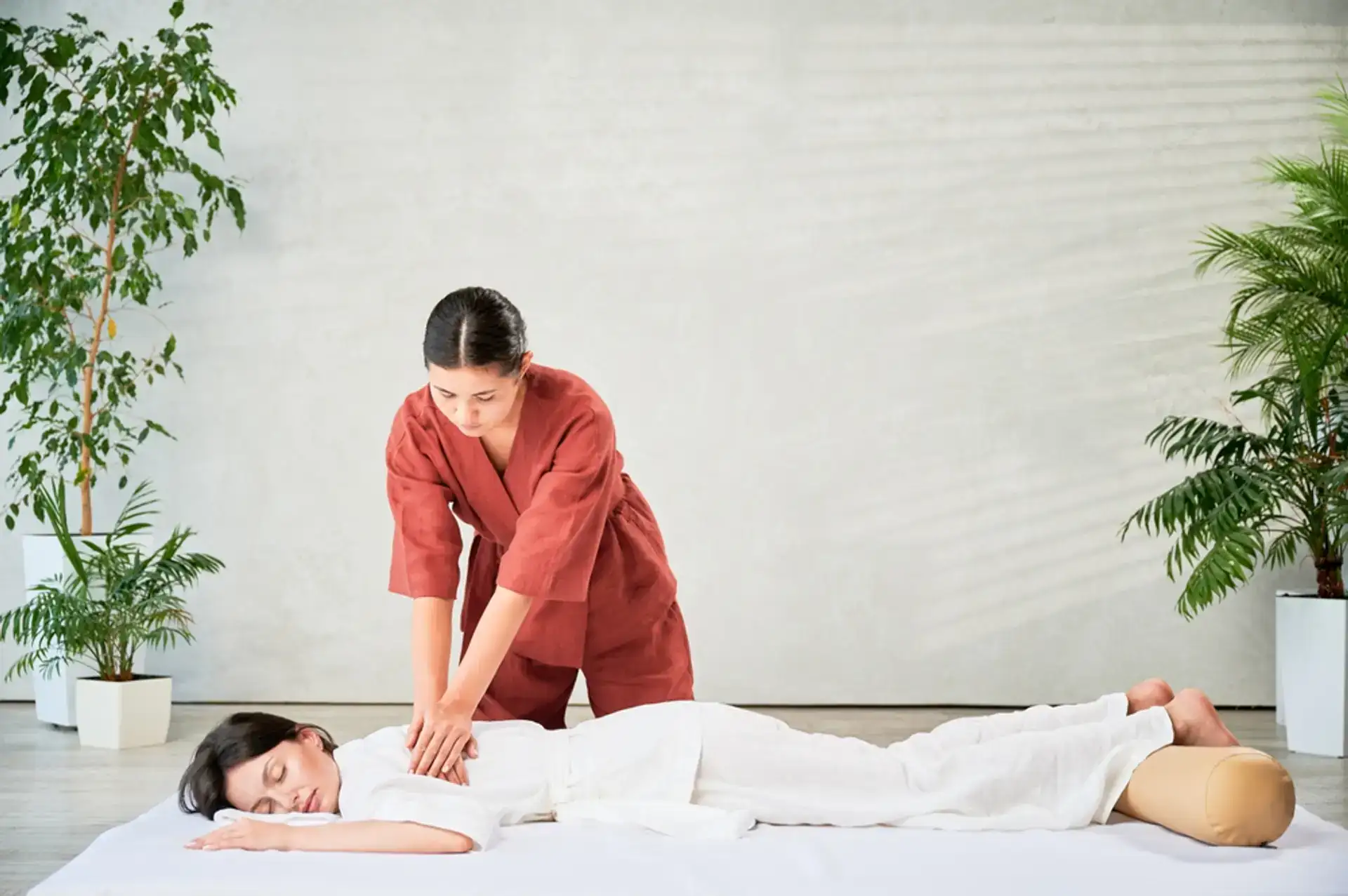A professional masseuse, dressed in a red outfit, is giving a back massage to a female client who is lying on her stomach. The massage is taking place indoors in a serene setting, likely a spa in Grand Rapids, MI.