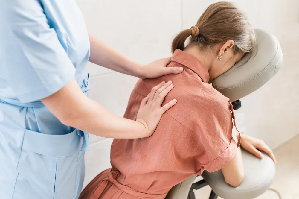 A woman sits on a massage chair with her face resting on the head support while a massage therapist in a blue uniform, offering chiropractor services in Grand Rapids, MI, massages her shoulders and upper back.