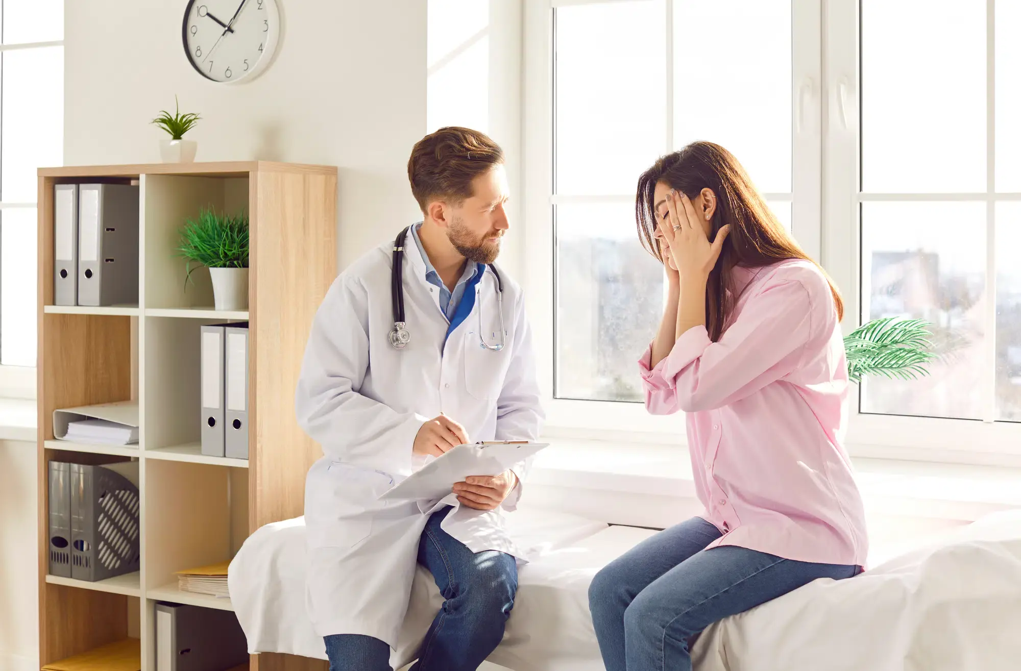A female patient sits on an examination table with her hands to her face, talking to a male doctor. She is expressing her symptoms and seeking headache pain relief at a clinic in **Grand Rapids, MI**.