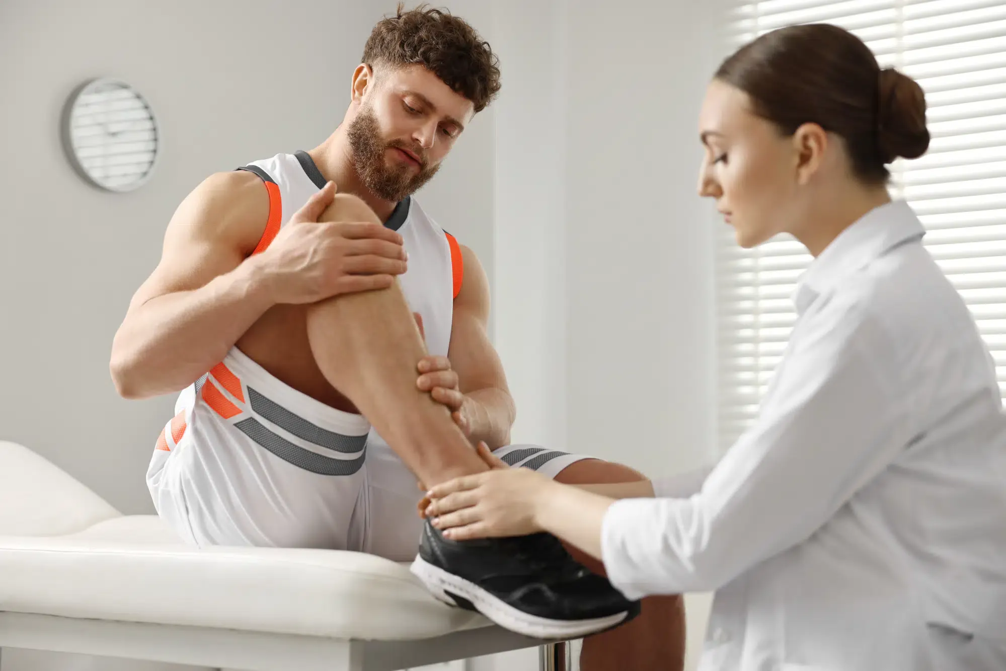 A female doctor examines a male athlete's leg and ankle on an exam table. The athlete is seeking treatment for a sports injury at a clinic in **Grand Rapids, MI