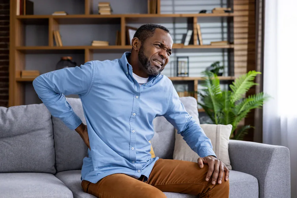 A man in Grand Rapids, MI, sits on a couch, grimacing and holding his lower back due to pain. He appears to be experiencing significant back discomfort.