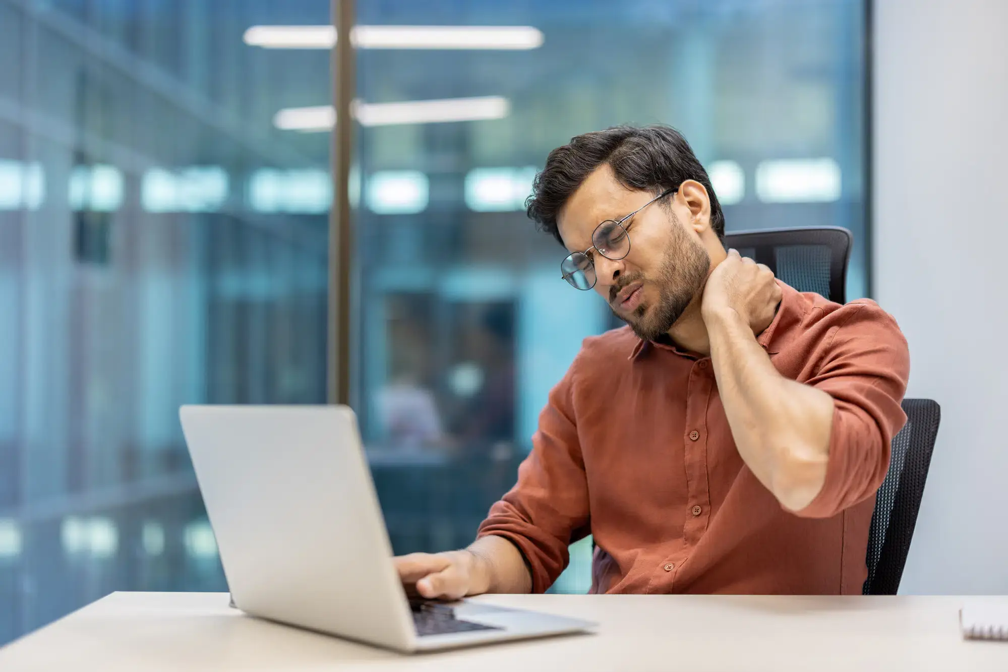 A man sits at his desk, wincing and holding his neck in pain while using his laptop. He may be suffering from neck pain and seeking chiropractic care for relief in **Grand Rapids, MI