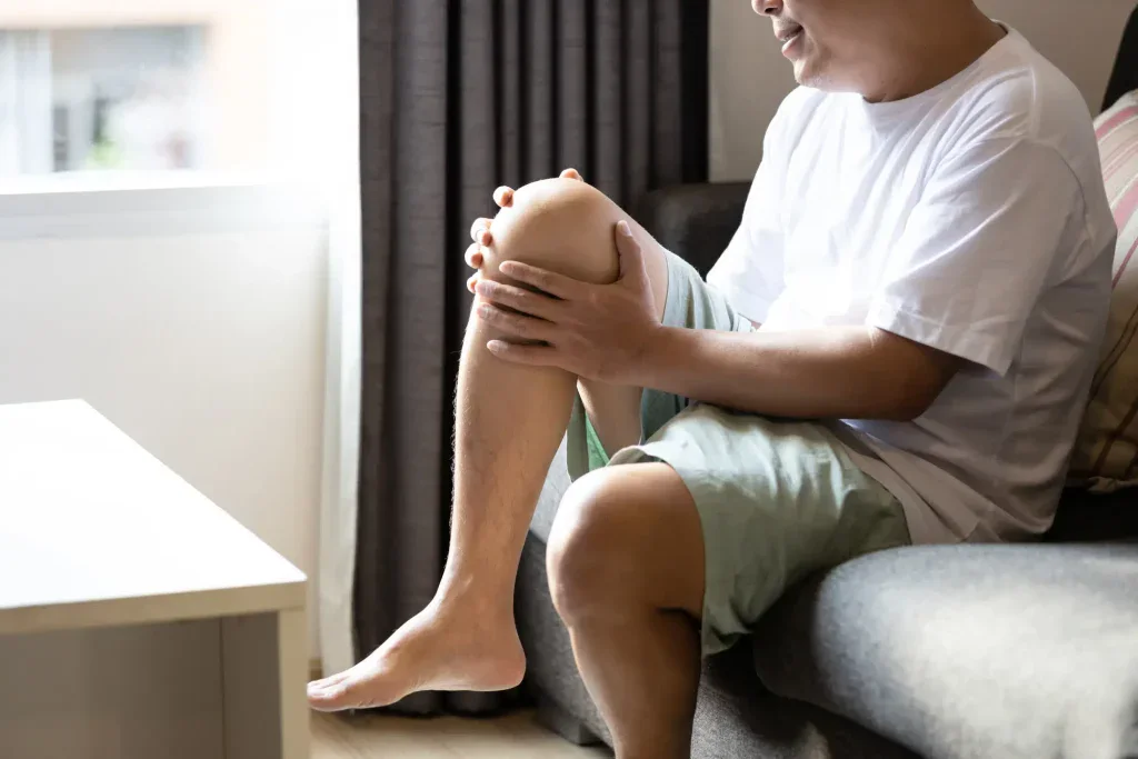 A man is sitting on a living room chair, clutching his knee to illustrate discomfort and leg pain. This scene depicts a common issue faced by residents in Grand Rapids, MI.