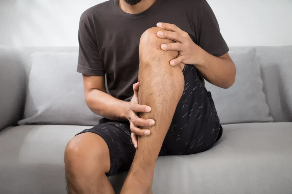 A man is sitting on a couch, holding his leg with a concerned expression due to pain. This image illustrates leg discomfort and pain in a home setting in Grand Rapids, MI.