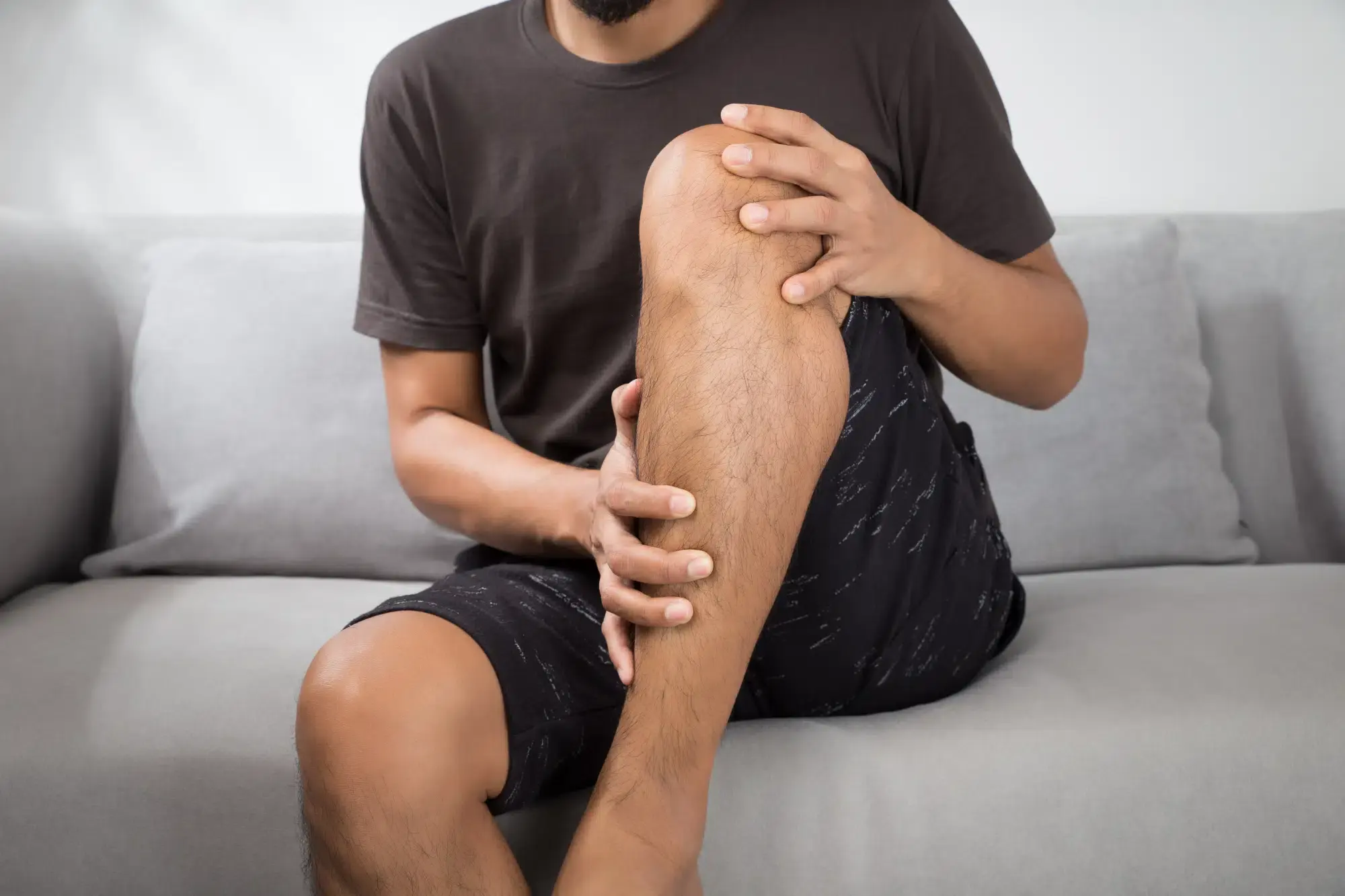 A man is sitting on a couch, holding his leg with a concerned expression due to pain. This image illustrates leg discomfort and pain in a home setting in Grand Rapids, MI.