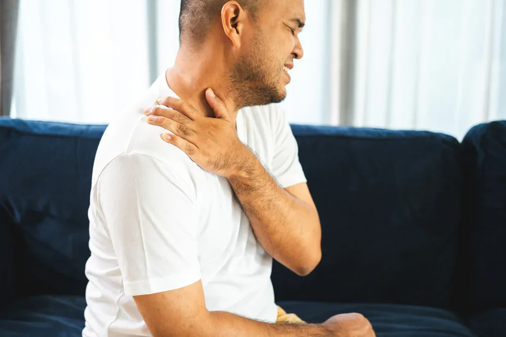 A man in a white t-shirt sits on a dark sofa, holding his shoulder and neck with a pained expression—highlighting discomfort that may benefit from Chiropractor Services Grand Rapids, MI. Light curtains are visible in the background.