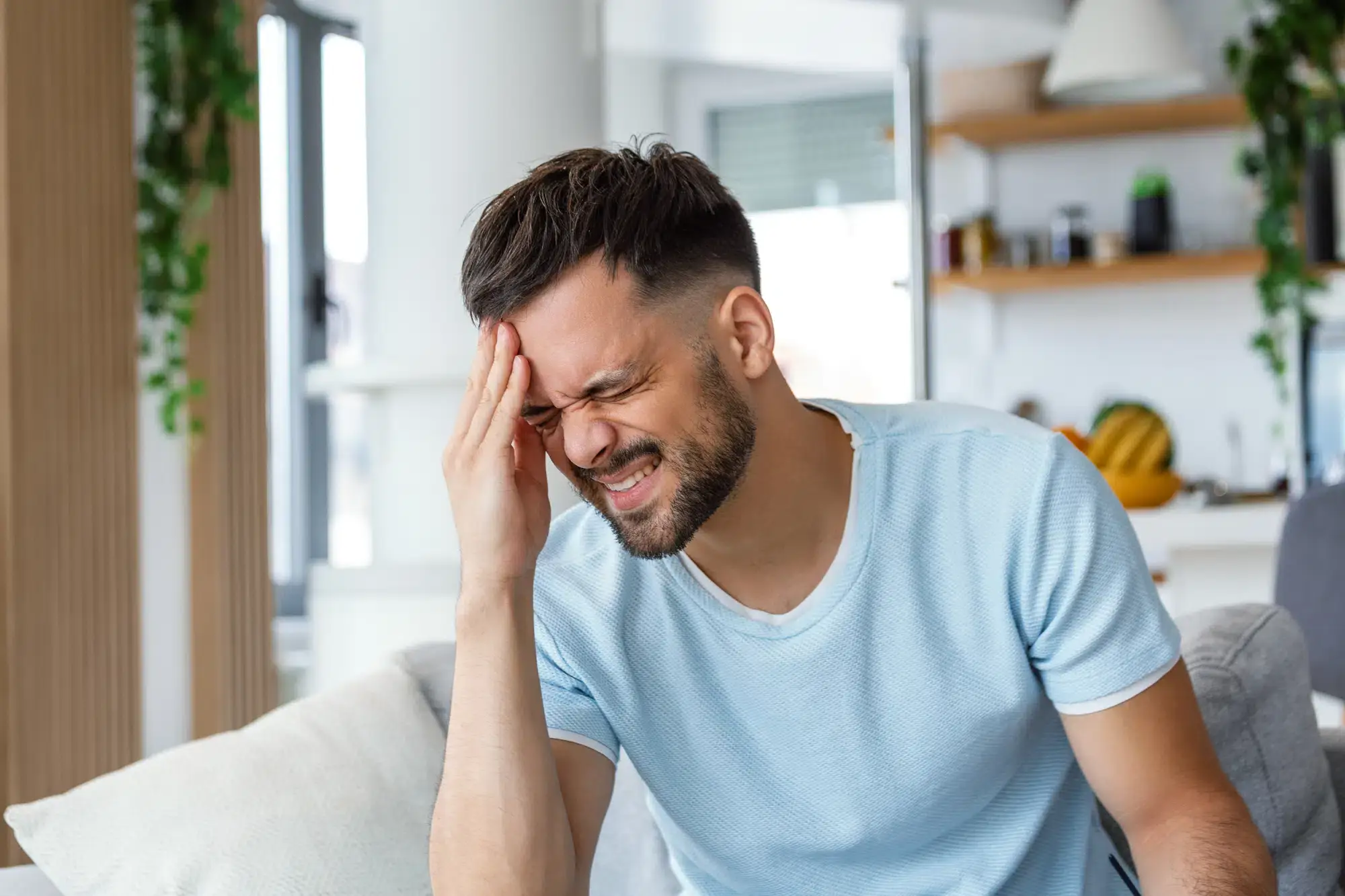 A person sits on a couch in a modern Grand Rapids, MI home, holding their head with one hand and eyes closed, surrounded by shelves, plants, and kitchen items—illustrating a moment of stress or headache.