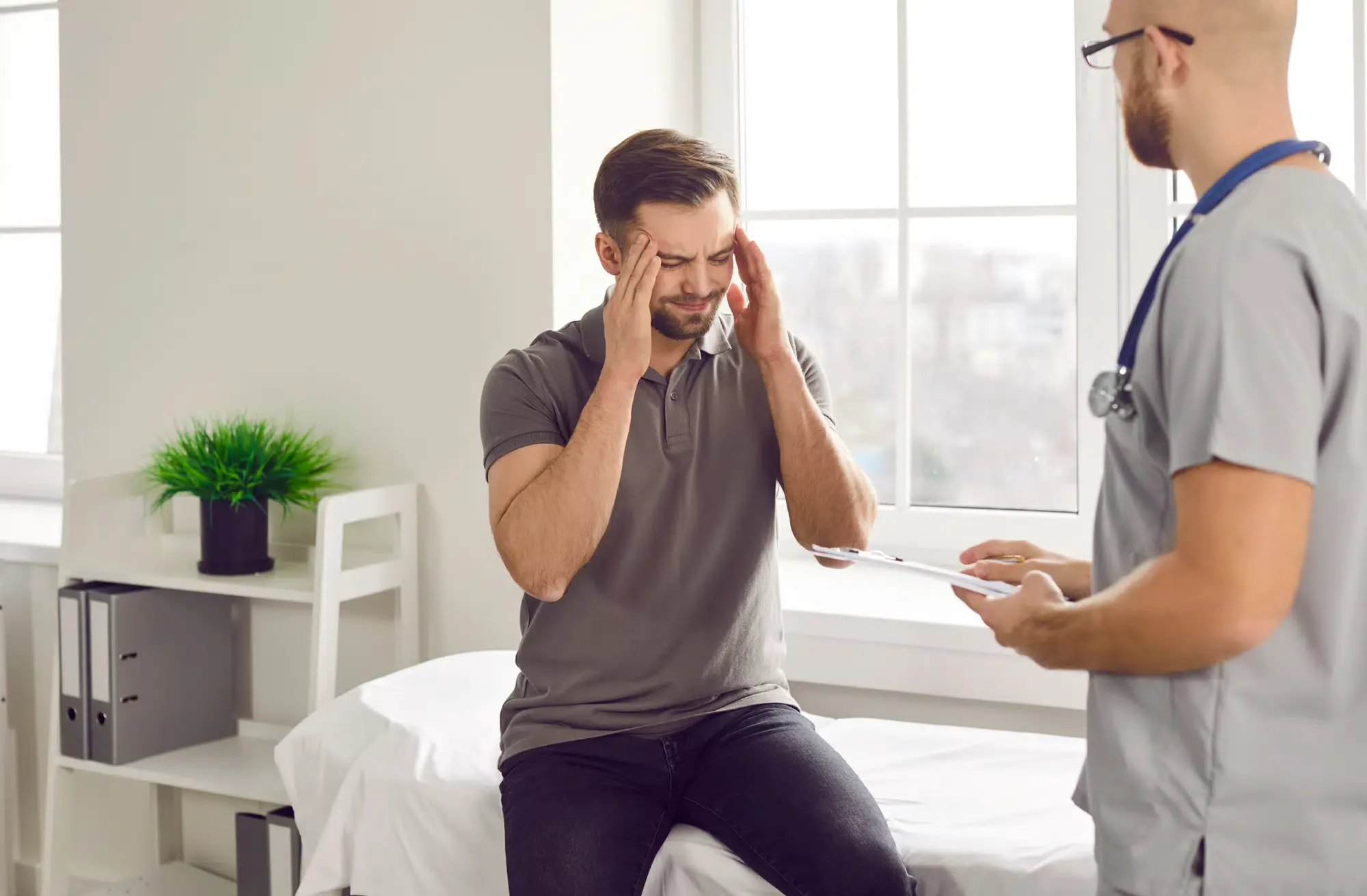 A male patient with his eyes closed and hands on his head is talking to a doctor about his headache. He may be seeking headache pain relief at a clinic in Grand Rapids, MI.