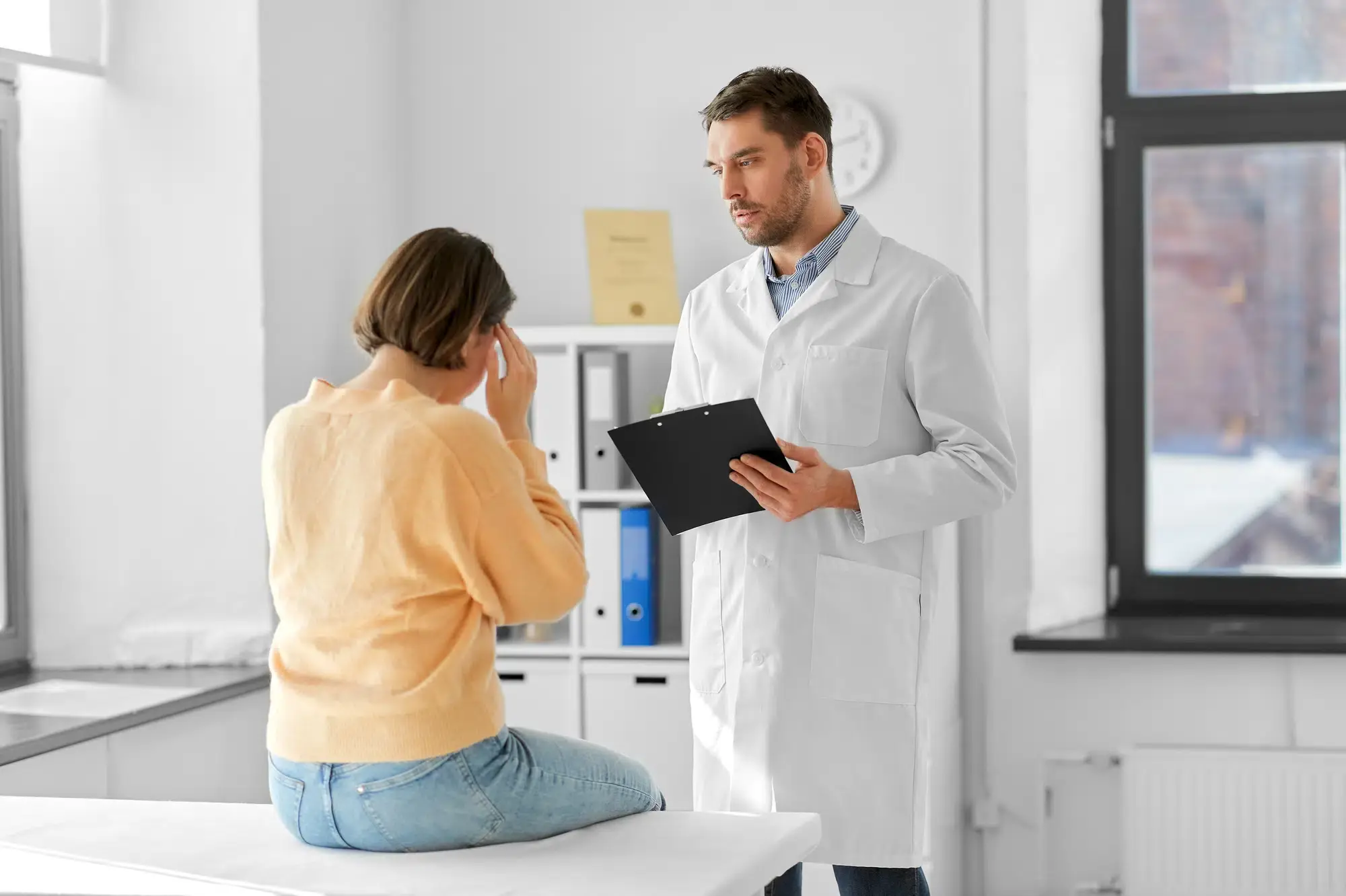 A young woman with her hand on her head speaks to a doctor in a clinic about her headache. She may be seeking migraine or headache pain relief in Grand Rapids, MI.