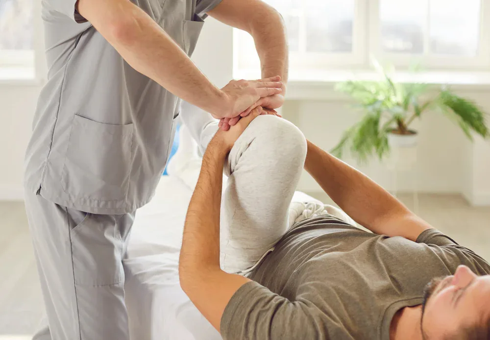 A physical therapist in medical scrubs performs a knee stretch on a patient lying on a treatment table in a bright room with large windows and indoor plants, illustrating a physical therapy session in Grand Rapids, MI.