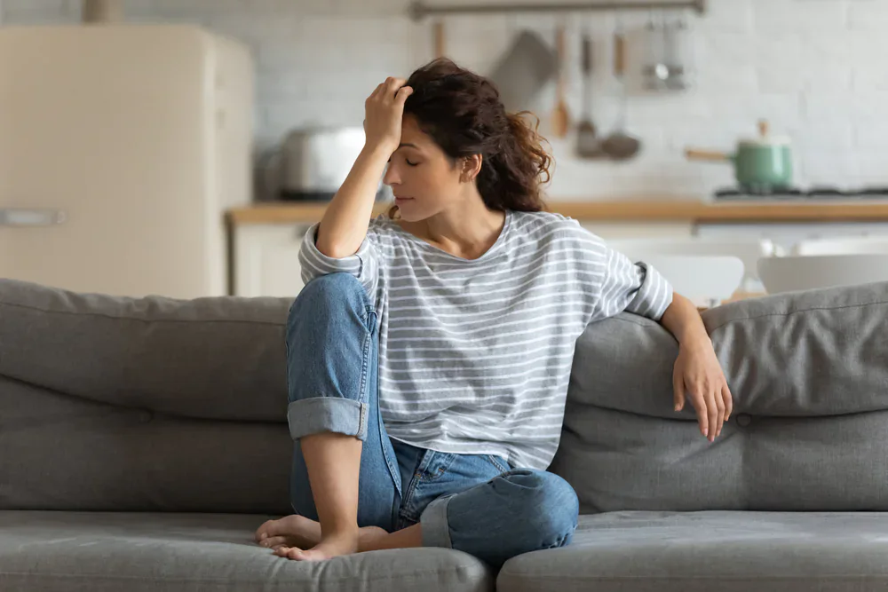 A person in a striped shirt and rolled-up jeans sits on a gray couch in a kitchen-living room space, with one hand on the couch and the other touching their head, surrounded by kitchen items like a stove, refrigerator, and hanging utensils—capturing a casual moment in a Grand Rapids, MI home.