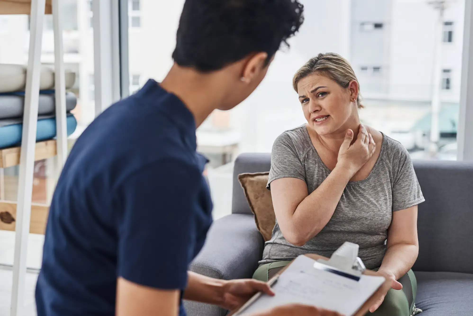A female patient sits on a couch, pointing to her neck while talking to a chiropractor. She is discussing her neck pain and seeking relief at a clinic in Grand Rapids, MI.