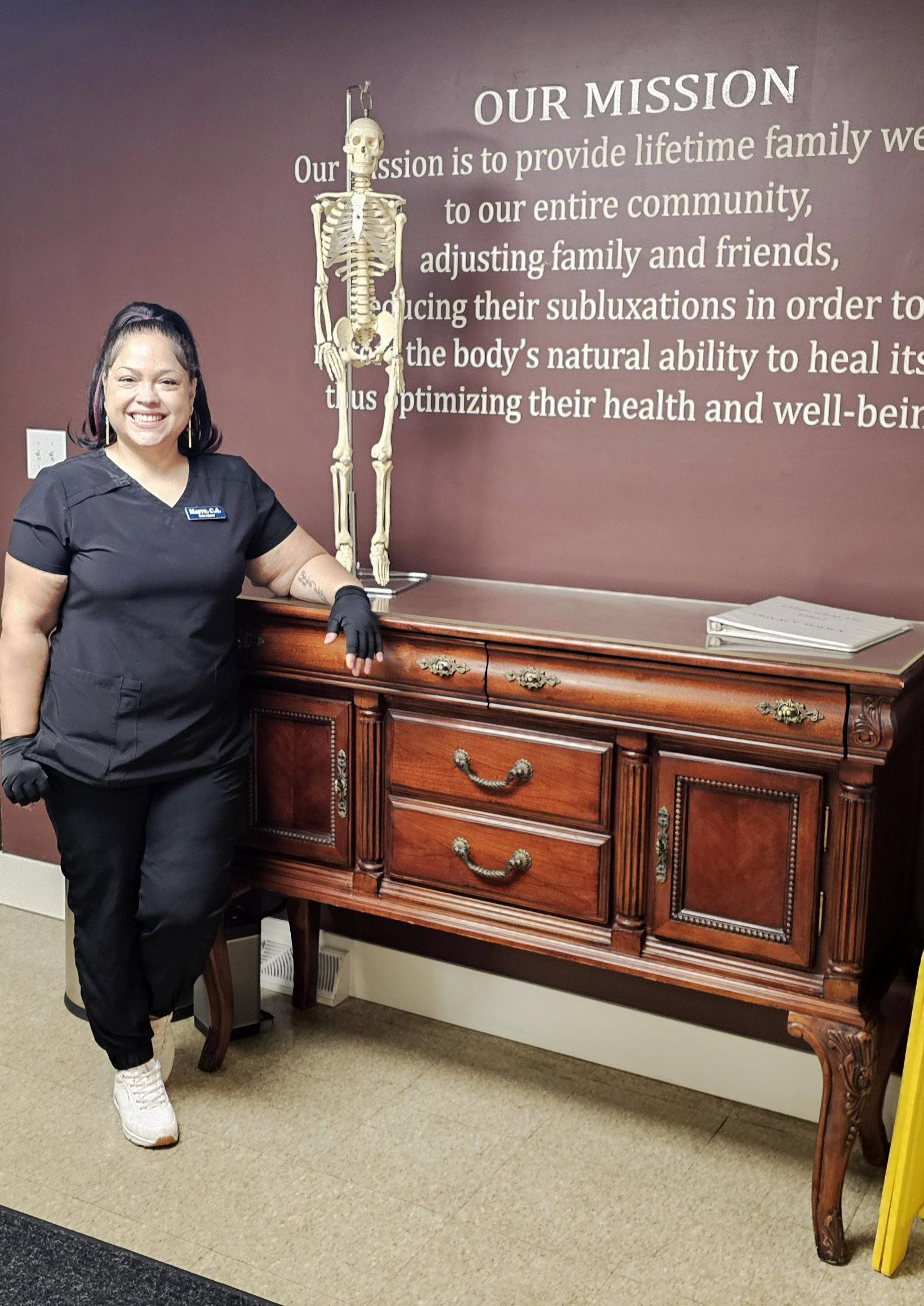 Chiropractic First staff member standing next to a wooden reception desk with a skeleton model in the background, emphasizing chiropractic care and holistic health services.