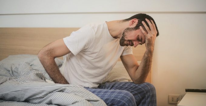 A man in Grand Rapids, MI, sits on the edge of a bed, holding his lower back in discomfort. He appears to be suffering from a headache, likely due to poor posture.