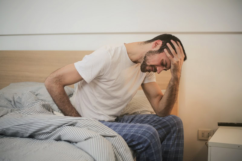 A man in Grand Rapids, MI, sits on the edge of a bed, holding his lower back in discomfort. He appears to be suffering from a headache, likely due to poor posture.