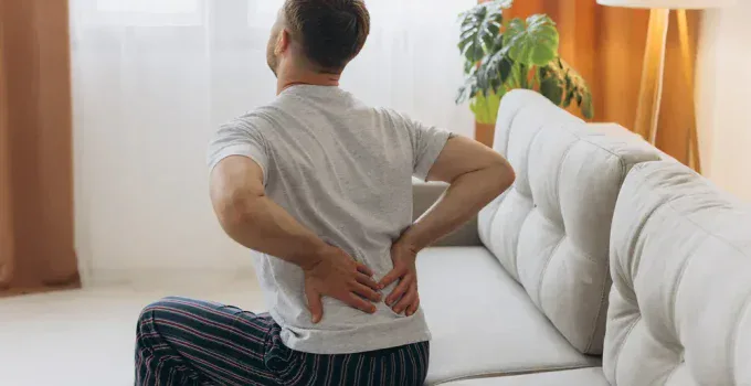 A man sits on a couch in MI, facing away, wearing striped pajama pants and a gray t-shirt. He holds his lower back with both hands in apparent pain as natural light filters through the window—perhaps in need of Chiropractor Services Grand Rapids.