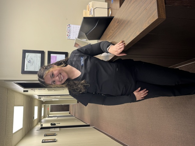 Woman in black scrubs smiles at a wooden reception desk with the Chiropractic Team in the hallway.